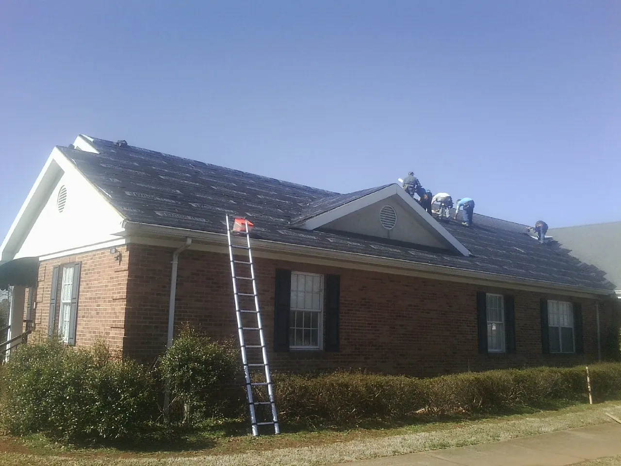 Crew tearing shingles off a brick ranch roof with a ladder set — Metal Roof Repair work by our crew in Sand Creek