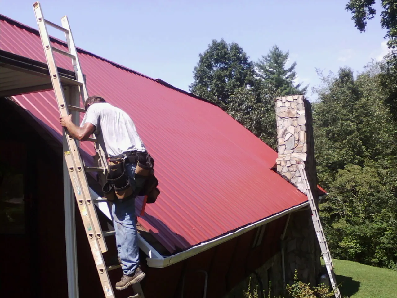 Roofer on a ladder beside a red metal roof and stone chimney — Soffit & Fascia Repair by Roofing Brothers in Sand Creek